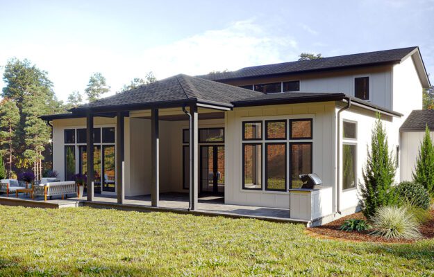 A modern, single-story home nestled in a lush, wooded landscape. The house features a large, covered porch with comfortable outdoor seating, expansive windows, and a well-manicured lawn surrounded by mature trees and shrubs, creating a serene and inviting outdoor living space.