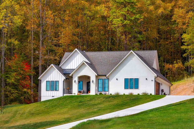 A modern, white farmhouse-style home nestled in a lush, autumnal forest. The house features a gabled roof, blue-framed windows, and a welcoming front porch. The surrounding landscape is ablaze with vibrant fall foliage, creating a picturesque and serene setting.