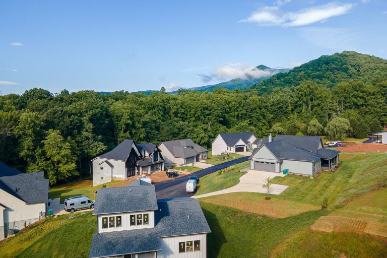 Aerial view of a residential neighborhood nestled in a lush, forested landscape. The image shows multiple modern houses with gabled roofs and attached garages, surrounded by well-manicured lawns and driveways. In the background, a densely wooded mountain range can be seen, with clouds partially obscuring the peaks. The overall scene conveys a peaceful, suburban setting in a picturesque natural environment.